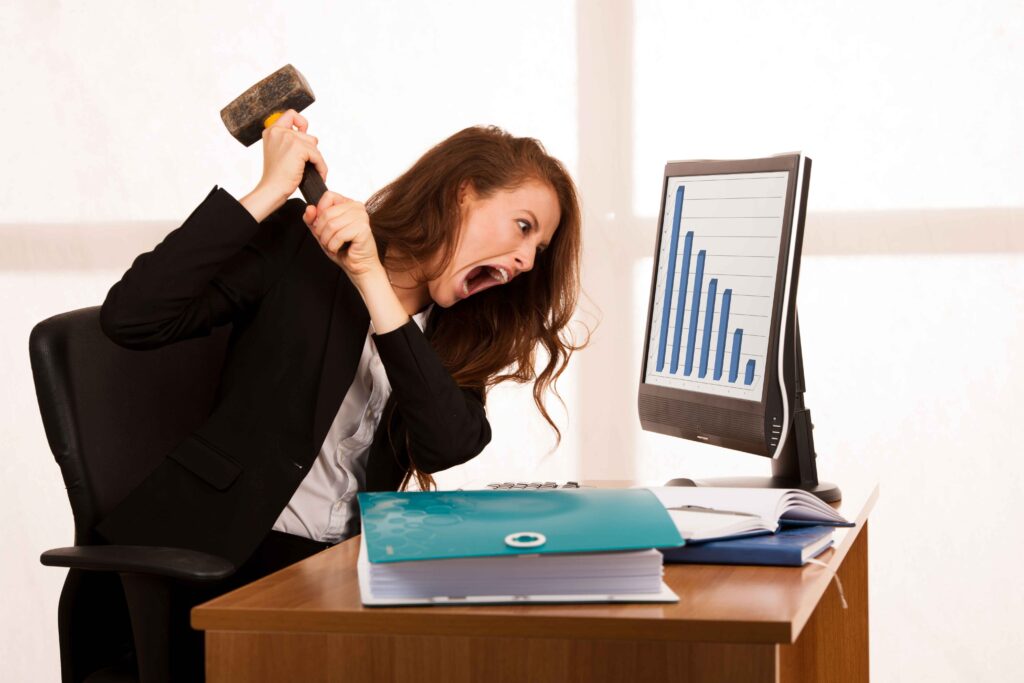 Hidden Costs of Staying Too Long in a Job You Hate - a woman at a desk with a hammer aimed at her desktop