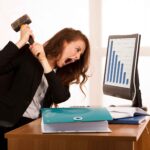 Hidden Costs of Staying Too Long in a Job You Hate - a woman at a desk with a hammer aimed at her desktop